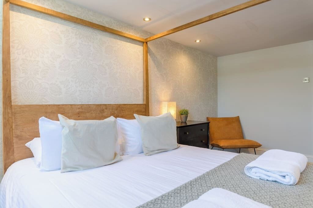 A neatly made bed with white linens and pillows, a small bedside table with a lamp, and a chair.