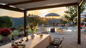 Un área de comedor al aire libre con una mesa preparada para dos personas, rodeada de sillas y sombrillas, con vistas a una piscina y montañas.