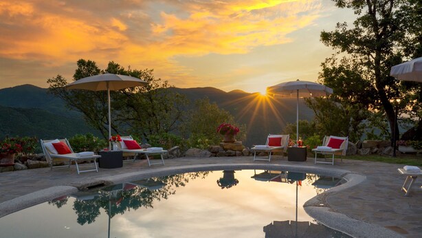 Zona de piscina con sillones reclinables y sombrillas, rodeada de montañas y un atardecer.