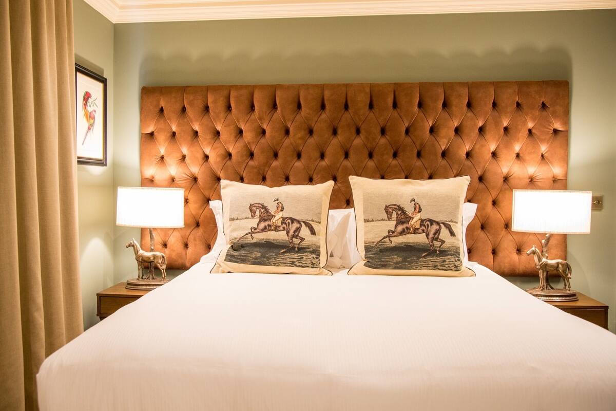 A hotel room with a tufted headboard, two matching pillows with horse designs, a nightstand with a lamp, and a framed picture on the wall.
