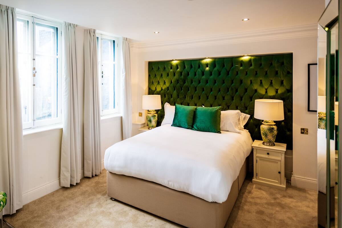 A hotel room with a large bed, two bedside lamps, a green upholstered headboard, and a view of the city through the window.