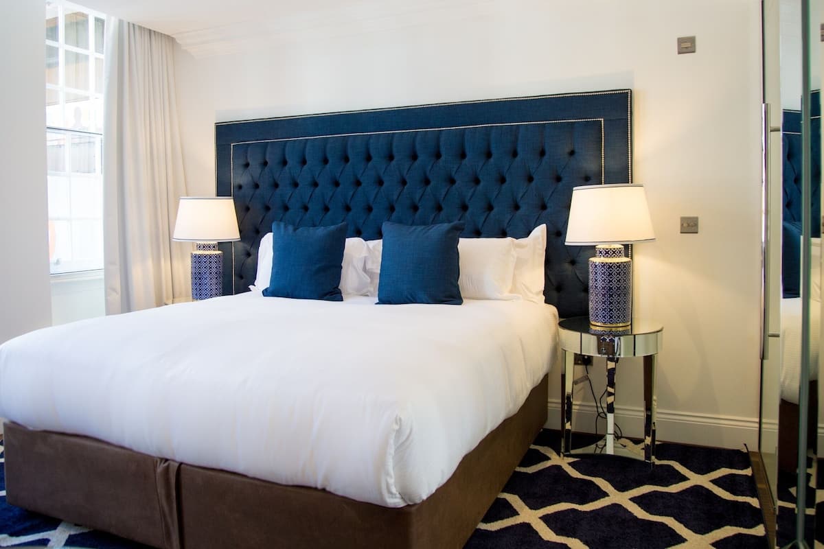 A large bed with a tufted headboard, two bedside lamps, and a patterned rug.
