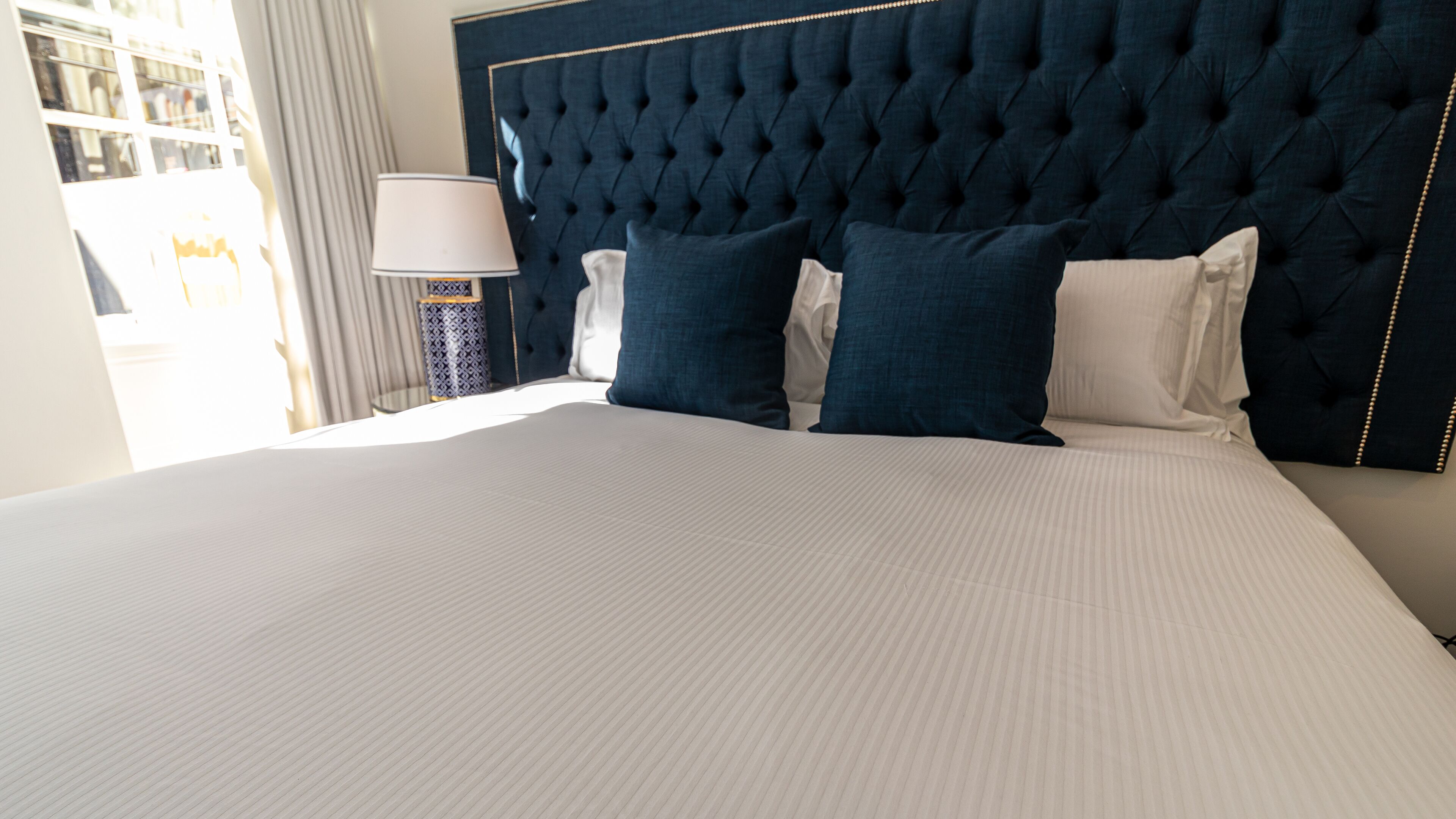 Una cama bien tendida con cabecera capitonada, una lámpara de noche y un jarrón estampado sobre una mesita de noche.
