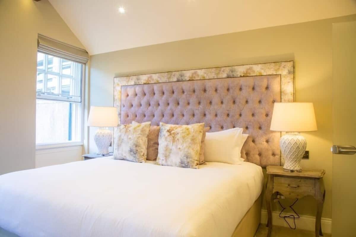 A large bed with a tufted headboard, two bedside lamps, and a window with blinds.