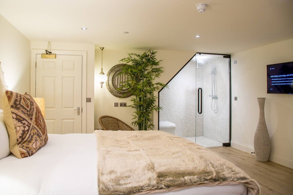 A bedroom with a bed, a shower, a wall-mounted TV, and a large plant.