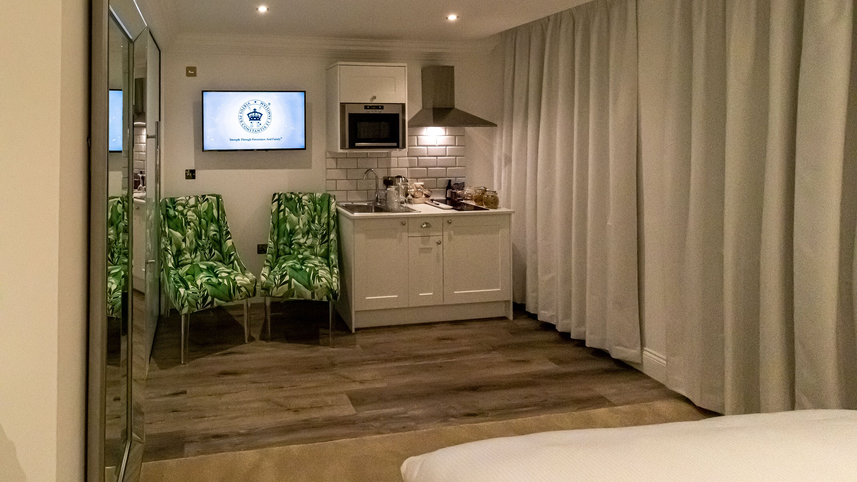 A hotel room with a bed, a TV mounted on the wall, a kitchenette with a microwave and oven, and a bathroom with a glass door.