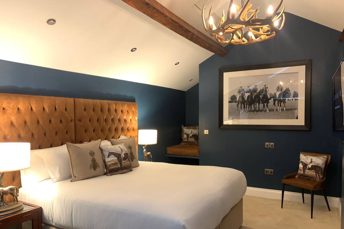 A bedroom with a large bed, a tufted headboard, a chandelier, a framed photograph on the wall, and a chair.