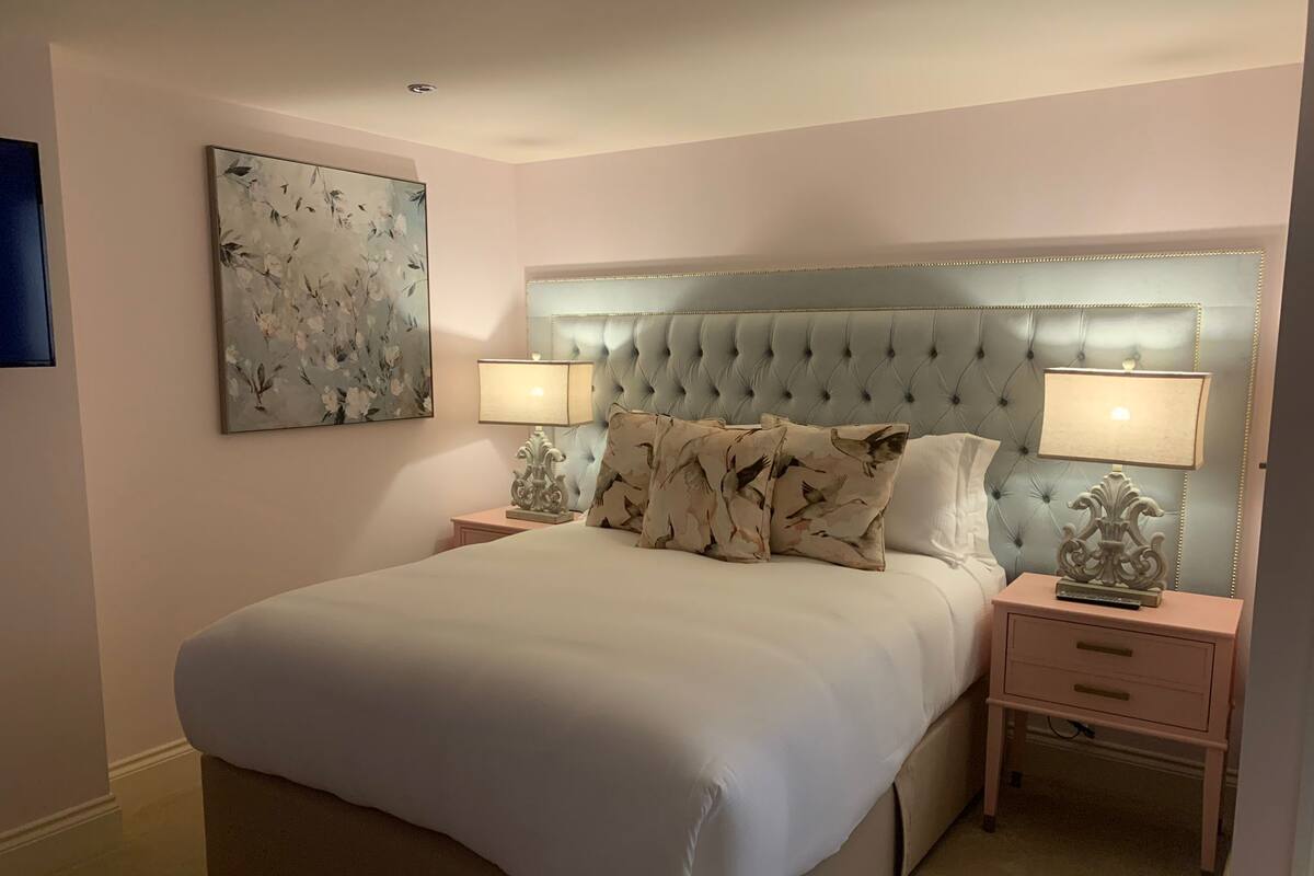 A bedroom with a tufted headboard, two bedside tables with lamps, a large bed, and a floral painting on the wall.