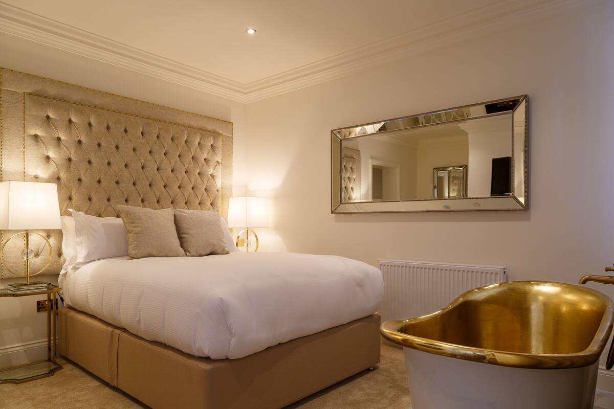 A bedroom with a large bed, two bedside lamps, a gold-framed mirror, and a gold bathtub.