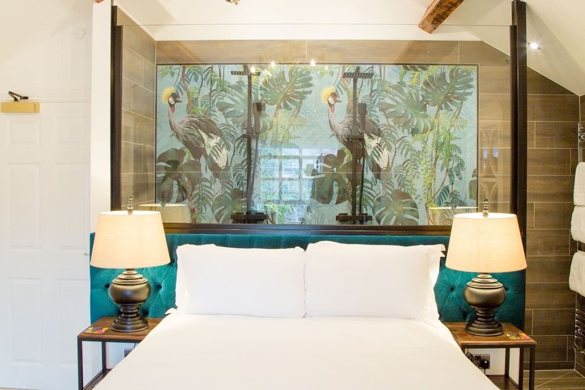 A bedroom with a large bed, two bedside tables with lamps, a chandelier, a large mirror, and a tropical-themed wall mural.