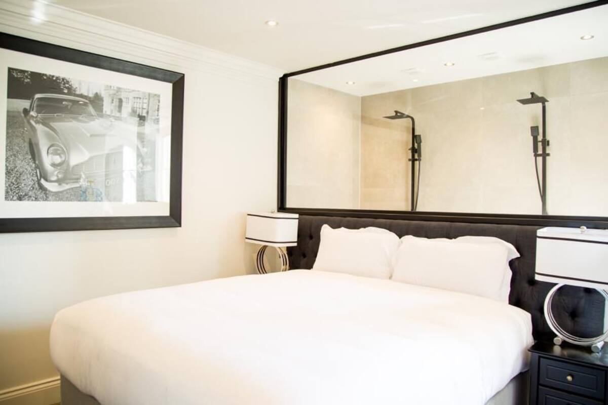 A modern hotel room with a large bed, a bedside table with a lamp, a large mirror, and a framed picture on the wall.