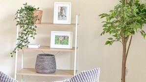 Una sala de estar con un televisor montado en la pared, un estante de madera con fotografías enmarcadas y una planta en maceta, y un árbol grande en maceta.