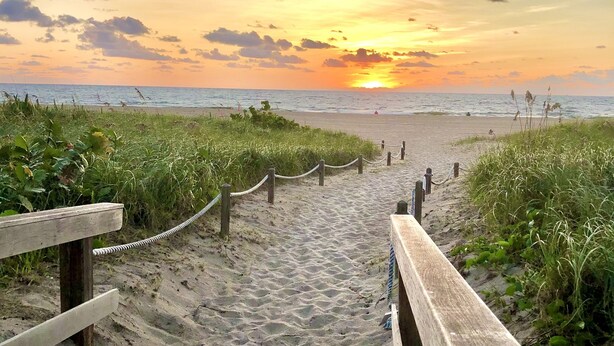 Ein hölzerner Steg führt zu einem Strand, im Hintergrund ist ein Sonnenuntergang zu sehen.