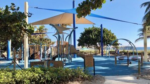 Ein Spielplatz mit verschiedenen Spielgeräten, darunter Rutschen und Kletterbereiche, beschattet durch große Sonnensegel.