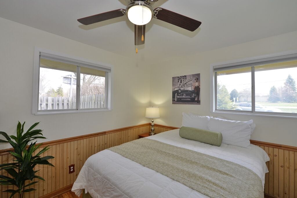 Ein Schlafzimmer mit einem Bett, einem Deckenventilator, zwei Fenstern, einem Nachttisch, einer Lampe und einem gerahmten Bild an der Wand.