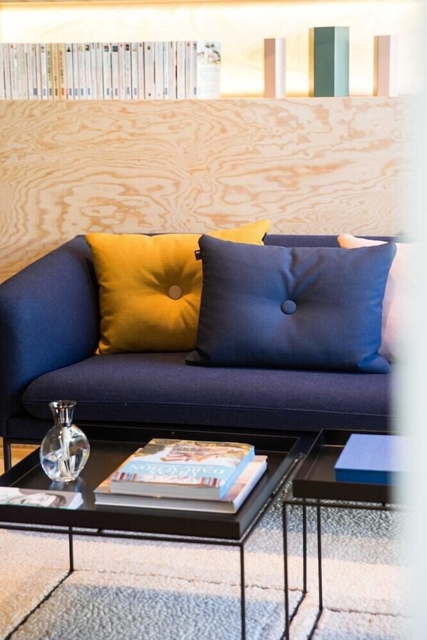 Ein modernes Wohnzimmer mit einem dunkelblauen Sofa, gelb-blauen Kissen, einem Glastisch mit Büchern und einem Dekoelement sowie einer Holzwand mit Bücherregal.