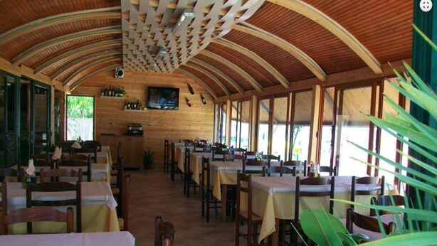 A restaurant with wooden walls and ceiling, tables with yellow tablecloths, and a TV screen on the wall. - Denta Hotel (Orikum)