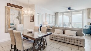 Un salon spacieux avec un grand canapé, une table à manger et des chaises, ainsi qu’un lustre. Une grande fenêtre offre une vue sur l’océan.