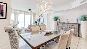 Une salle à manger moderne avec une table à plateau en verre, des chaises recouvertes d’un tissu beige, un luminaire décoratif au plafond et une vue sur le salon grâce à de grandes fenêtres.