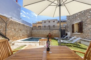 A wooden table with two wine glasses and a bottle, a stone wall, a swimming pool, and lounge chairs in a backyard setting.