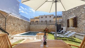 Una taula de fusta amb dues copes de vi i una ampolla, una paret de pedra, una piscina i gandules en un jardí.