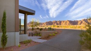 Un bâtiment moderne doté d’un patio, offrant une vue dégagée sur un paysage désertique avec des montagnes en arrière-plan, et une allée pavée menant à l’entrée.