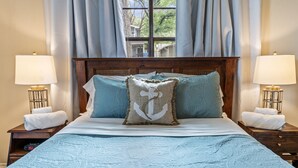 A neatly made bed with a blue bedspread and anchor-themed pillows, flanked by two bedside tables with lamps and folded towels. - Lakefront w/ Dock, 300mb WIFI, Theater, Arcade (Tool)