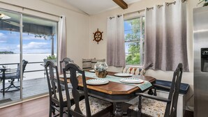 A dining area with a table set for four, chairs with floral upholstery, a ship's wheel decoration, and a view of the water through large windows. - Lakefront w/ Dock, 300mb WIFI, Theater, Arcade (Tool)