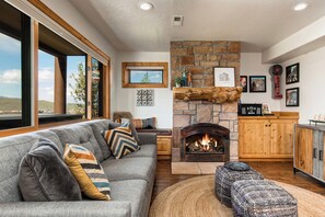 A cozy living room with a stone fireplace, a grey sofa with patterned cushions, and a view of the outdoors.