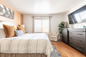 Un dormitorio con cama, televisor, cómoda, silla y ventana con cortinas.