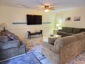 A living room with a sofa, a bench, a coffee table, a TV, a ceiling fan, and a patterned rug.