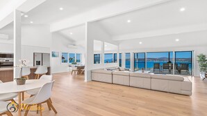 A spacious living area with a large sofa, dining table, and chairs, a kitchen area, and a balcony with a view of the sea. - Randall Drive No 64 - Magnificent Views - Pool - Air con - WiFi - The list goes on! (Salamander Bay)