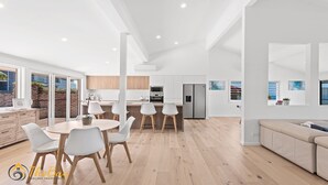 A modern kitchen with white cabinets, a stainless steel refrigerator, and a dining area with a round table and white chairs. - Randall Drive No 64 - Magnificent Views - Pool - Air con - WiFi - The list goes on! (Salamander Bay)