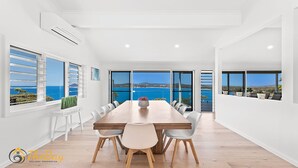 A spacious dining area with a long wooden table, white chairs, and a view of the sea through large windows. - Randall Drive No 64 - Magnificent Views - Pool - Air con - WiFi - The list goes on! (Salamander Bay)