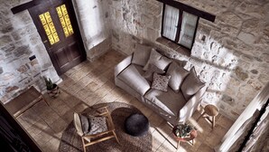 Une pièce rustique avec un mur en pierre, une porte en bois ornée de motifs jaunes, un canapé beige agrémenté de coussins à motifs, une chaise en osier et une petite table en bois sur laquelle est posé un bol rempli de plantes.