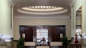 Ein Hotel-Empfangsbereich mit rotem Teppich, zwei Zimmerpflanzen, einer Rezeptionistin und einem hölzernen Empfangstresen.