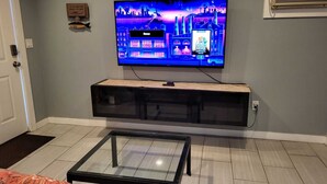 Una sala de estar con un televisor de pantalla plana montado en la pared, una mesa de centro de vidrio y una puerta con felpudo.