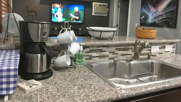 Una cocina con un lavamanos de acero inoxidable, una cafetera y un microondas. Hay dos televisores montados en la pared, una cesta sobre la encimera y una puerta que da a otra habitación.