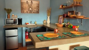 Una cocina funcional con microondas, refrigerador y lavamanos. Hay dos taburetes verdes y una mesa con un plato y un vaso de cobre. Un cuadro de un ciervo adorna la pared, y estantes con diversos objetos están sobre la encimera.