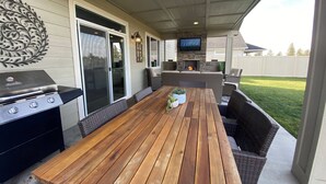 An outdoor patio with a wooden table, wicker seating, a grill, and a TV mounted on a stone wall. - Beautiful 3 bed+den, 3bath huge home on the Spokane River and Centennial Trail (Spokane Valley)