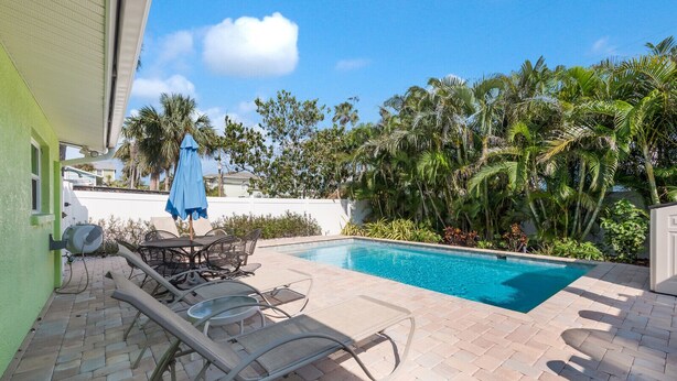 A swimming pool with lounge chairs and a table, surrounded by palm trees and a green building. - 2 Bedroom / 2 Bath Ground Level With a Heated Pool (Holmes Beach)