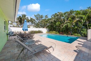 A swimming pool with lounge chairs and a table, surrounded by palm trees and a green building.