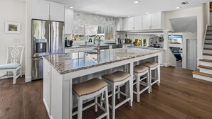 Une cuisine moderne avec des meubles blancs, un grand îlot central doté de tabourets, des appareils électroménagers en acier inoxydable et une vue sur l’extérieur.