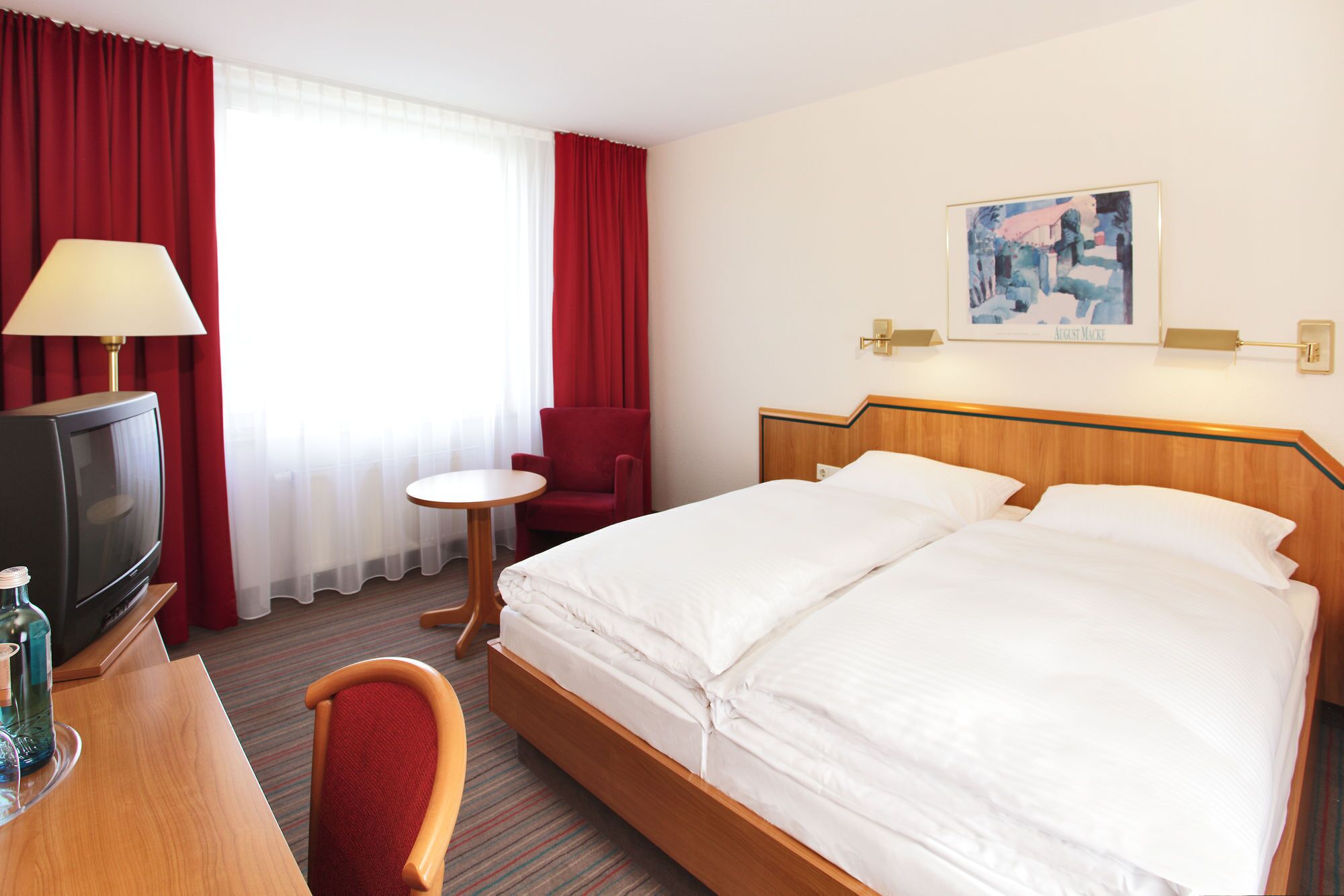 A hotel room with a bed, a desk with a lamp, a chair, a television, and a painting on the wall.