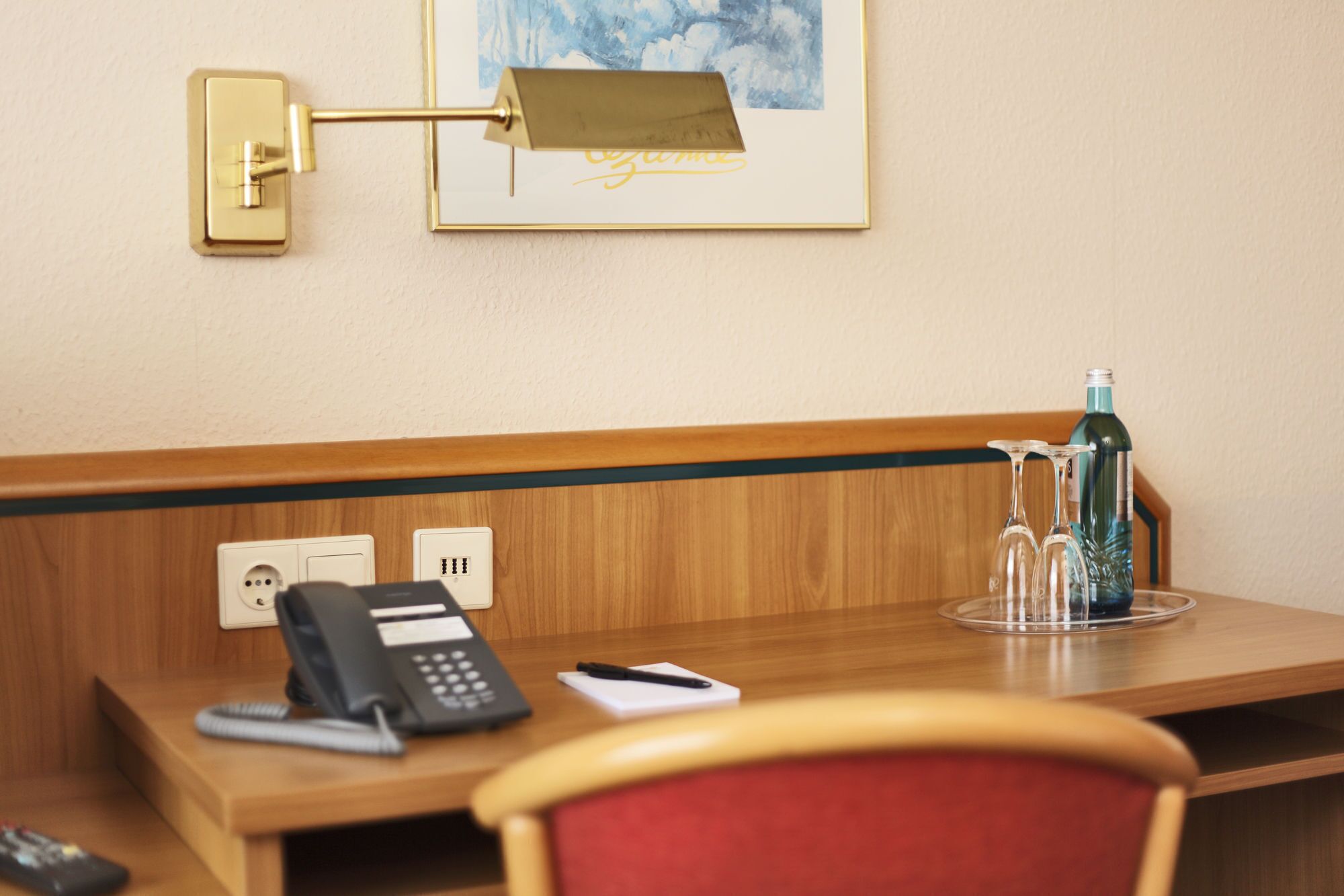 A hotel room with a wooden desk, a telephone, a notepad, a pen, a remote control, and a framed picture on the wall.