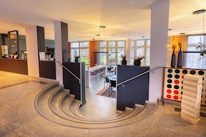 A modern hotel lobby with a staircase, a reception desk, and a dining area.