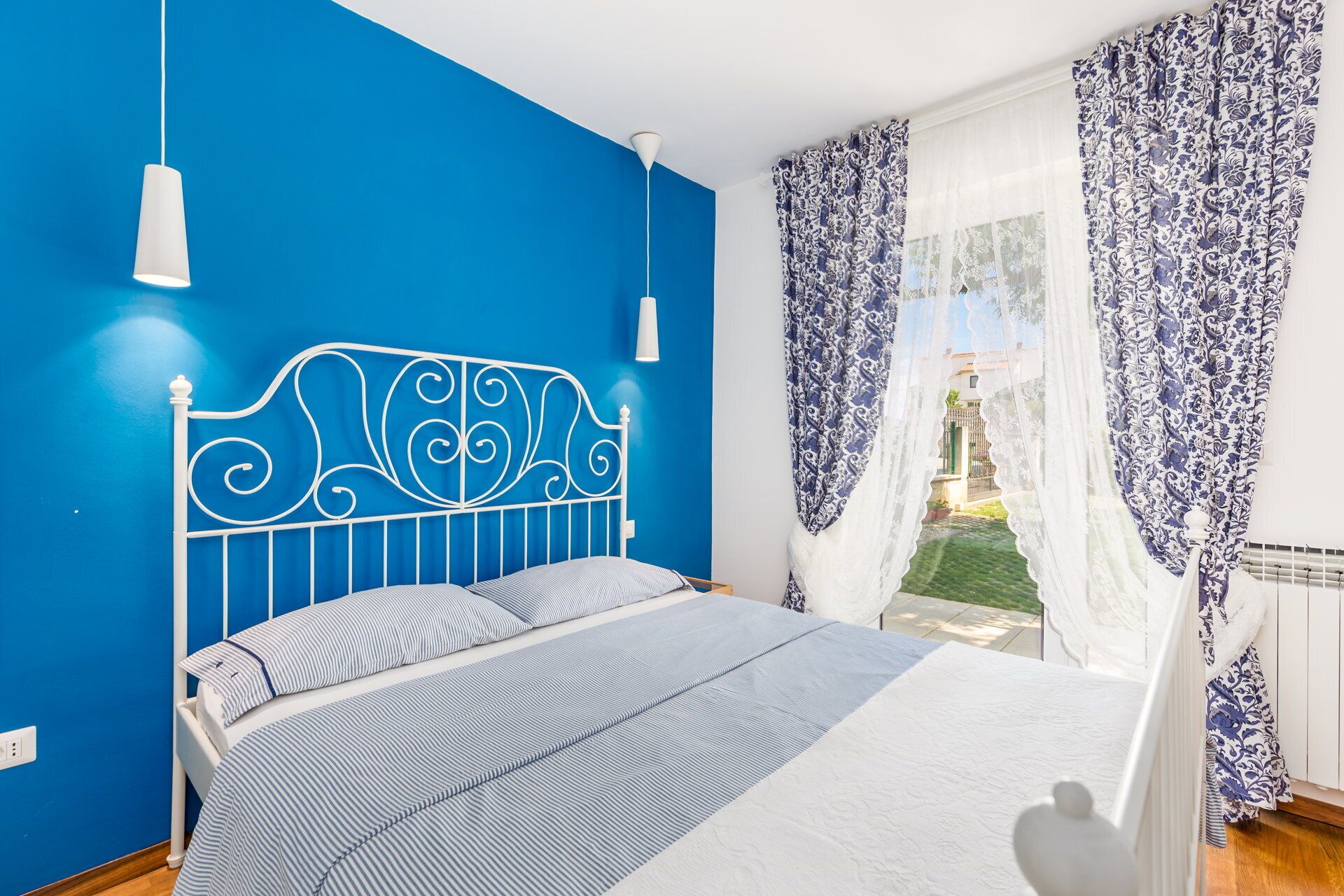 Camera da letto con parete blu, testiera in metallo bianco, letto con biancheria a righe e vista su un giardino attraverso tende trasparenti.