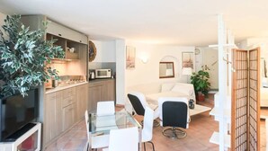 A modern living room with a glass dining table, white sofas, and a kitchen area with wooden cabinets and a microwave.