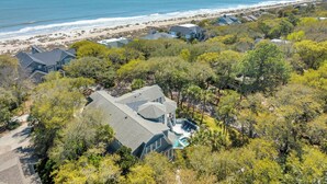 A coastal property with a large house featuring a swimming pool, surrounded by dense trees and a beach in the background.