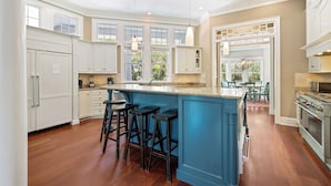 A kitchen with wooden flooring, white cabinets, a blue island with bar stools, and a view to a dining area.